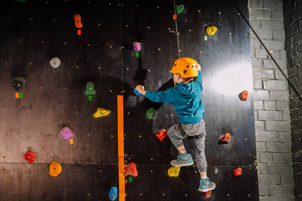 Valmiera, Latvia - March 28, 2025 - Young boy in a yellow helmet climbs an indoor rock wall, using colorful grips while secured with a harness and rope, focused on reaching the top.