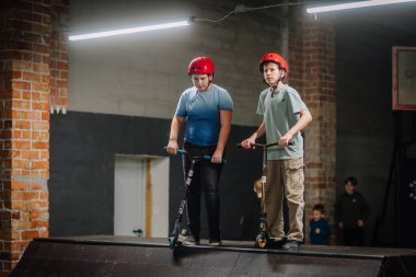 Valmiera, Letonya - 28 Mart 2025 - Kırmızı kasklı iki çocuk arka planda diğer çocuklar oynarken kapalı bir kaykay parkında mobiletlerin üzerinde dikiliyorlar..