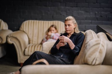 Valmiera, Letonya - 28 Mart 2025 - bej renkli bir kanepede oturan genç bir kadın akıllı telefon tutuyor, odaklanmış görünüyor. Arka planda bir çocuk, rahat bir salonda bir fincan ile sessizce oturur..