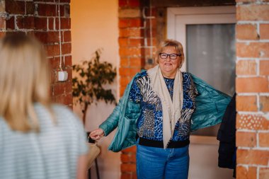 Valmiera, Letonya - 28 Mart 2025 - Gözlüklü ve renkli bir bluz giyen gülümseyen yaşlı kadın, elinde bir çanta ve palto tutarak evin içinde, çerçevenin dışında biri tarafından karşılanıyor..