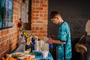 Valmiera, Letonya - 28 Mart 2025 - Mavi gömlekli bir çocuk, içecekler, atıştırmalıklar ve kağıt bardaklarla dolu bir parti masasında soda şişesi açıyor. Etkinlik, tuğla duvar arka planıyla birlikte kapalı mekanda ayarlandı.
