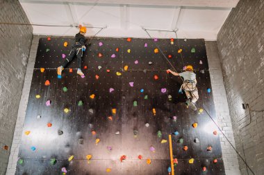 Valmiera, Letonya - 28 Mart 2025 - Kask ve koşum takımı takan iki çocuk güvenlik halatları kullanarak renkli bir kaya duvarına tırmanıyor, eğlenceli ve aktif bir eğlence yarışmasına katılıyorlar..