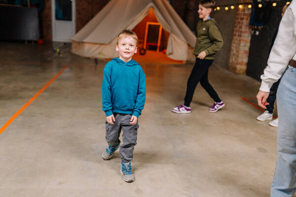 Valmiera, Latvia - March 28, 2025 - Young boy in a teal hoodie stands on an indoor concrete floor, smiling toward the camera. A tent and other kids are visible in the background.