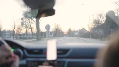 Valmiera, Letonya - 2 Mart 2025 - Gün batımında sakin bir banliyö sokağında, arka planda ağaçlar ve evler bulunan bir otobüs durağından geçen bir arabanın içine bakın..