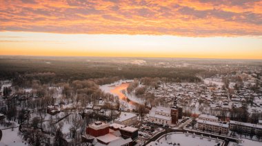 Valmiera, Letonya - 12 Ocak 2022 - Gün batımında karlı bir kasabanın gökyüzü manzarası parlayan bir nehir çevreyi sararken, yoğun bir orman ve yukarıda sıcak turuncu bulutlar.