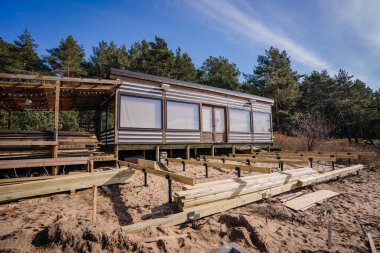 Geniş pencereli ahşap bir kulübe ve bitişik bir teras, açık ve aydınlık bir günde, bir çam ormanının yakınındaki kumlu bir alanda yapım aşamasında..
