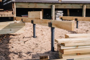 Güverte inşaatı sırasında tahta kirişlere ve destek direklerine yakın çekim, kumlu zeminde istiflenmiş tahtalar ve arka planda ahşap bir bina.