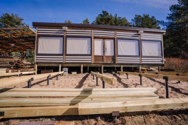 Çağdaş ahşap kulübe, kumlu topraklarda temel kirişleri olan, ormanla çevrili, açık ve güneşli bir günde cepheden izlenen..