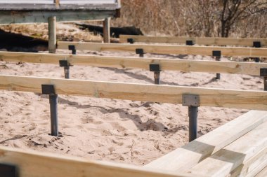 Metal destekli kumlu toprakta, ahşap güverte iskeleti yapısına yakın çekim. Açık hava ortamındaki bir kulübeye yakın yapısal ilerlemeyi gösteriyor..