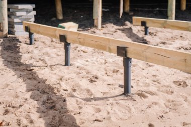 Güverte inşaatı sırasında metal direklere monte edilmiş ahşap kiriş, kumlu toprakta, kaldırım blokları yakın ve ahşap bir yapının bir parçası görünür.