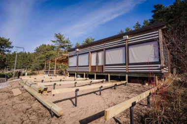 Kumlu zeminde ahşap terası olan ormandaki modern kulübe, güneşli bir günde metal destekleri ve kereste kirişleri döşenmiş..