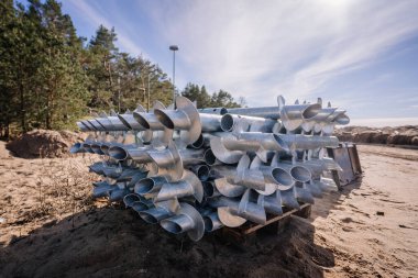 Ormanın yakınındaki kumlu bir inşaat sahasında, parlak bir gökyüzünün altında kurulmaya hazır, bir paletin üzerine yığılmış metal vida yığını..