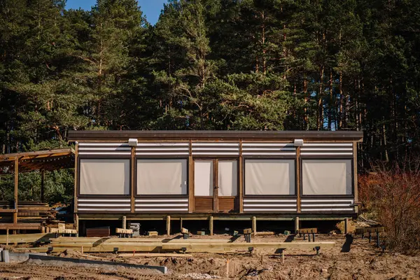 Kapalı jaluzili çizgili ahşap bir kulübe, arka planda inşaat malzemeleri ve orman ağaçları bulunan kumlu arazinin ortasındaki cambaz ayaklıklarında yükseliyor..