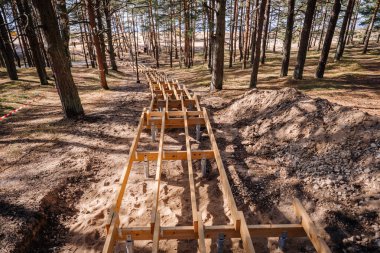 Tahta merdiven iskeleti kumlu bir orman tepesinde, ağaçlar ve dikkat bantlarıyla çevrili, uzaktaki sahile doğru gidiyor..