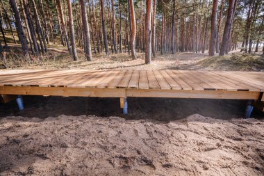 Çam ormanının kumlu zemininde yeni inşa edilmiş ahşap bir kaldırım. Zemin vida temelleriyle destekleniyor. Güneş ışığı güvertede gölgeler bırakıyor..