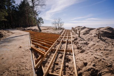 Tahta kaldırım iskeleti yapım aşamasında. Orman ve sahil kenarında kumlu bir patika. Uzakta gökyüzü ve okyanus görünüyor..
