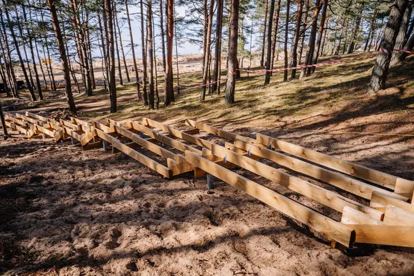 Çam ormanındaki kumlu bir yamaçta inşaat halindeki ahşap merdiven korkulukları, metal zemin çapalarıyla destekleniyor ve güvenlik bandıyla çevrili..