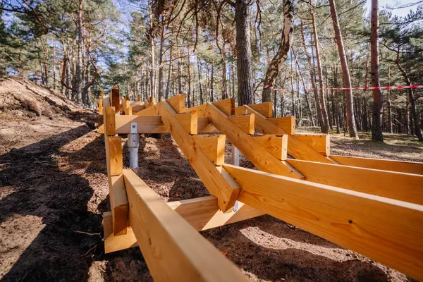Orman yamacında inşaat halindeki ahşap bir merdivenin yakın görüntüsü. Metal direklerle destekleniyor. Çam ağaçlarının arasından süzülen güneş ışığı..