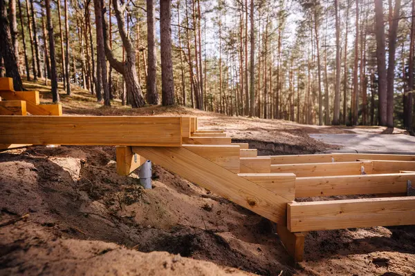 Ormanda yapım aşamasındaki ahşap merdivenler, kumlu arazi üzerine inşa edilmiş metal vida yığını temelleri kullanılarak, yüksek çam ağaçları ve doğal ışık ile çevrelenmiş..