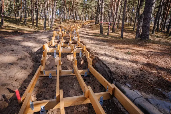 Kum ve çam ağaçlarıyla çevrili görünür vida yığını temelleri ve metal parantezleri ile bir ormanda yapım aşamasındaki ahşap iskelet..