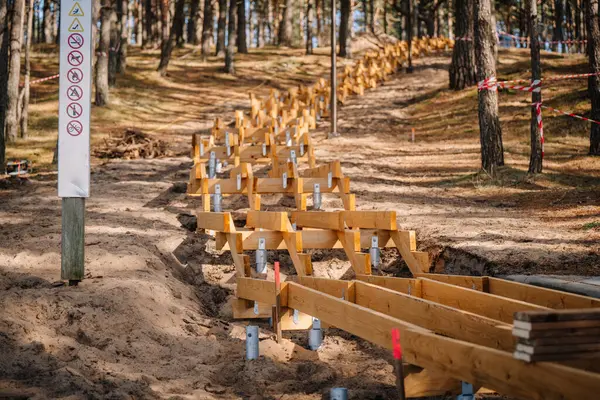 Görünür metal vida yığınları, destek kirişleri ve kumlu yol boyunca güvenlik uyarısı olan bir ormanda yapım aşamasındaki tahta merdiven..
