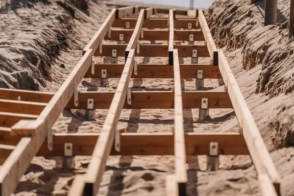 Metal çapalar ve parantezlerle desteklenen kumlu arazide yapım aşamasındaki ahşap kaldırım iskeleti, açık havada iki kumsalın arasına dizilmiş..