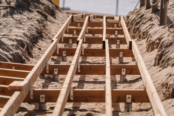 Metal çapalar ve parantezlerle desteklenen kumlu arazide yapım aşamasındaki ahşap kaldırım iskeleti, açık havada iki kumsalın arasına dizilmiş..