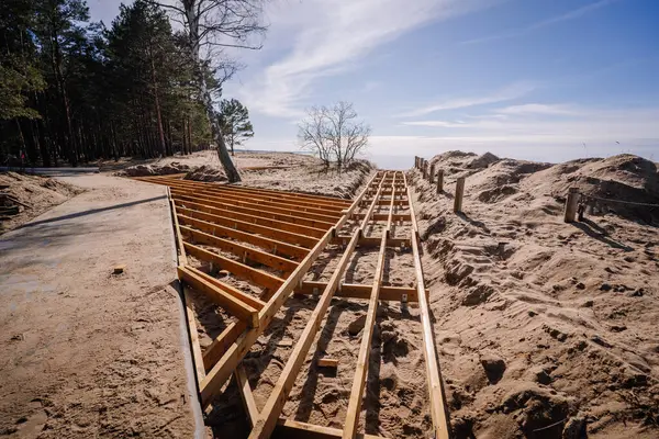 Tahta kaldırım iskeleti yapım aşamasında. Orman ve sahil kenarında kumlu bir patika. Uzakta gökyüzü ve okyanus görünüyor..