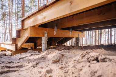 Orman bölgesindeki kumlu arazide metal vida yığınları tarafından desteklenen ahşap platform yapısı, alt güverte iskeleti ve metal parantezler sergileniyor.