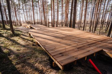 Çam ormanında yapım aşamasındaki geniş ahşap bir platform güvertesi, görünür bir çerçeve ve yapının yanında kırmızı bir kanal borusu var..