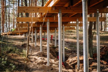 Bir sıra galvanizli çelik vida yığını bir orman bölgesindeki ahşap bir platformu destekliyor ve düz olmayan kumlu arazi üzerinde hassas bir yapı gösteriyor..