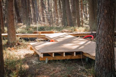 İnşaat rüzgarları altında, görünür iskeleti ve kırmızı borusu olan metal tornavida yığınlarıyla desteklenen bir orman boyunca ahşap bir tahta kaldırım..