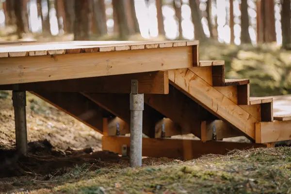 Ormandaki yüksek bir tahta kaldırıma bağlı yeni yapılmış ahşap bir merdivenin yakın çekimi. Çimenli arazi üzerindeki metal direklerle destekleniyor..