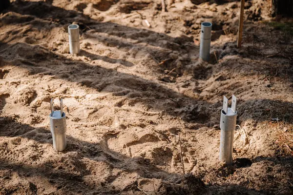 Bir inşaat sahasında kısmen kumlu toprağa monte edilmiş metal vidalar, ormanlık bir alanda temel çalışması için hazırlanmış..