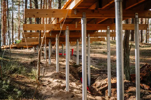 Bir sıra galvanizli çelik vida yığını bir orman bölgesindeki ahşap bir platformu destekliyor ve düz olmayan kumlu arazi üzerinde hassas bir yapı gösteriyor..