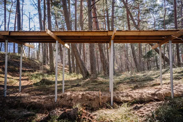 Yüksek ahşap kaldırım inşaat halinde. Ormanlık alanda kumlu arazi ve yüksek çam ağaçları olan metal vida yığınları destekliyor..