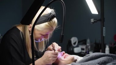Bir güzellik uzmanı parlak salon ışıkları altında kırbaç uzatma prosedürü uyguluyor. Müşteri tedavi sırasında battaniyeyle rahatça örtünüyor..