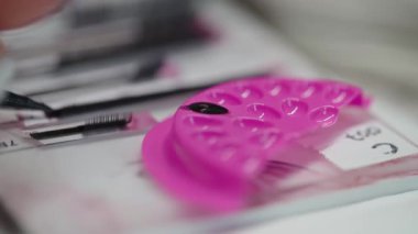 Profesyonel kirpik uygulama prosedürlerinde kullanılan pembe bir yapıştırıcı paletine siyah kirpik yapıştırıcı batıran bir cımbız videosu..