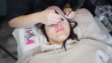 Güzellik teknisyeni, güzellik salonunda uzanan bir kadına kirpik takar. Profesyonel güzellik ortamında parlak ışık altında hassas aletler kullanır..