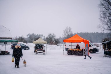 Valmiera, Letonya - 6 Şubat 2022 - Karla kaplı zemin, çadırlar ve mal satan satıcılarla küçük bir kış pazarı. Kırsal bir ortamda, kar altında köpeğini dolaştıran bir kadın..