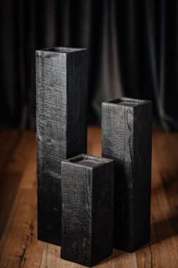 Ahşap zeminde, koyu arkaplan ile farklı yüksekliklerde üç dikey yanık tahta blok. Kömürleşmiş doku ve çatlaklar açıkça görülüyor..