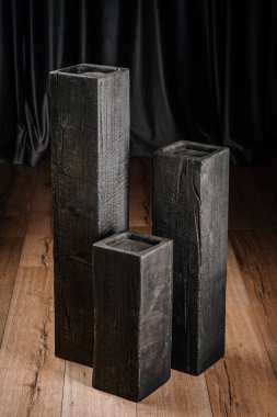 Ahşap zeminde, koyu arkaplan ile farklı yüksekliklerde üç dikey yanık tahta blok. Kömürleşmiş doku ve çatlaklar açıkça görülüyor..