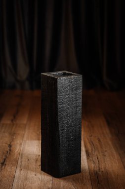 Kömürleşmiş ahşap bir vazo ya da pürüzlü bir dokusu olan içi boş bir blok, arka planda koyu perdeleri olan ahşap bir zeminde duruyor. Ahşap temasını yak.