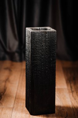 Kömürleşmiş ahşap bir vazo ya da pürüzlü bir dokusu olan içi boş bir blok, arka planda koyu perdeleri olan ahşap bir zeminde duruyor. Ahşap temasını yak.