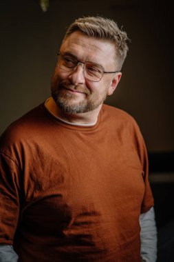 Valmiera, Letonya - 12 Şubat 2022 - Gözlüklü ve kahverengi gömlekli gülümseyen bir adam sıcak iç aydınlatma ile yan tarafa bakar. Portre, rahat ve düşünceli bir ifadeyi yansıtıyor..