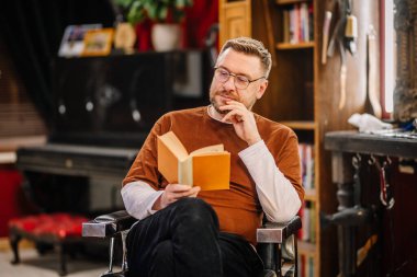 Valmiera, Letonya - 12 Şubat 2022 - Gözlüklü ve sakallı bir adam rahat bir odada otururken kitap okur. Sakin ve odaklanmış görünüyor, çenesini dikkatlice tutuyor..