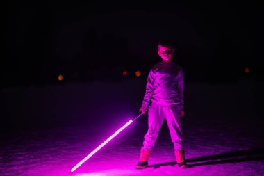 Gece vakti, elinde parlak mor bir ışın kılıcı tutan çocuk, karanlık, kış ortamında neon ışıklarıyla çarpıcı bir şekilde aydınlatılıyor..