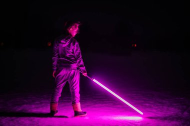 Bir çocuk gece elinde parlak mor bir ışın kılıcıyla karların üzerinde duruyor, çarpıcı bir şekilde neon ışıkla yüzünü çeviriyor ve sahneyi bilim kurgu tarzında aydınlatıyor..