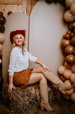 Madona, Letonya - 15 Şubat 2025 - Kırmızı şapkalı ve süet çizmeli gülen kadın, balon ve özel tabela ile batı temalı bir kutlamada saman balyasında poz veriyor..