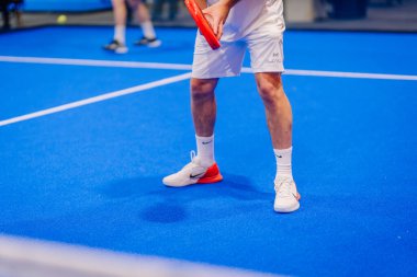Riga, Letonya - 5 Nisan 2025 - canlı mavi bir iç sahada elinde raket, beyaz spor giysisi ve Nike ayakkabıları olan bir erkek oyuncunun yakın çekimi.